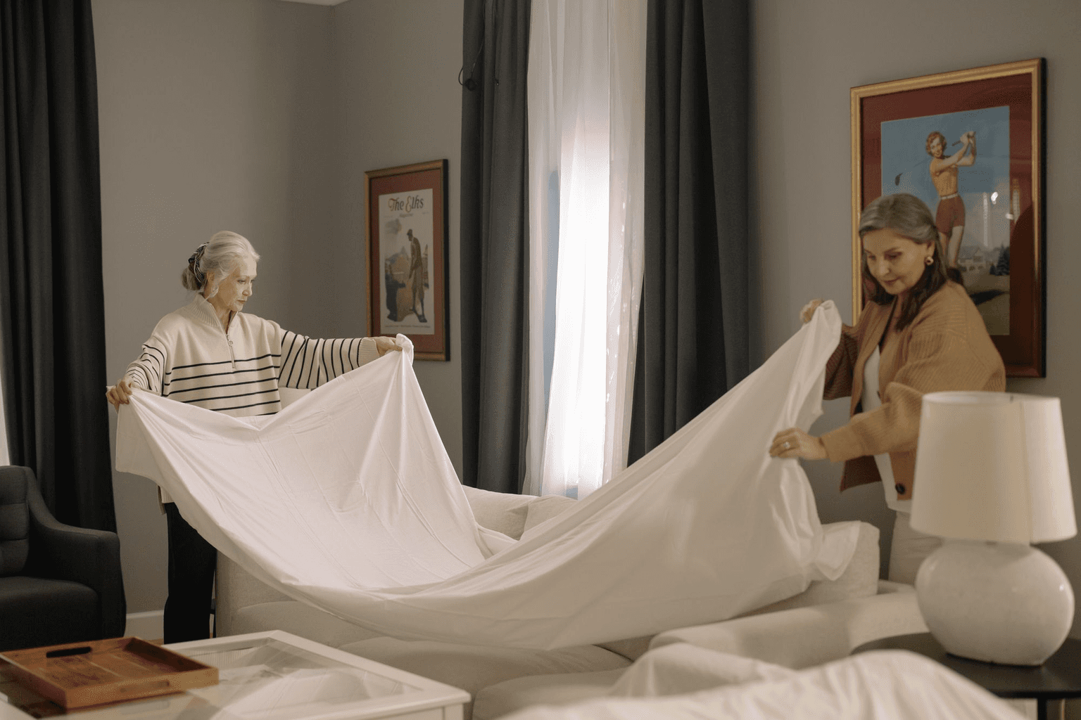 A younger woman and an older woman spreading a clean sheet together in her living room.