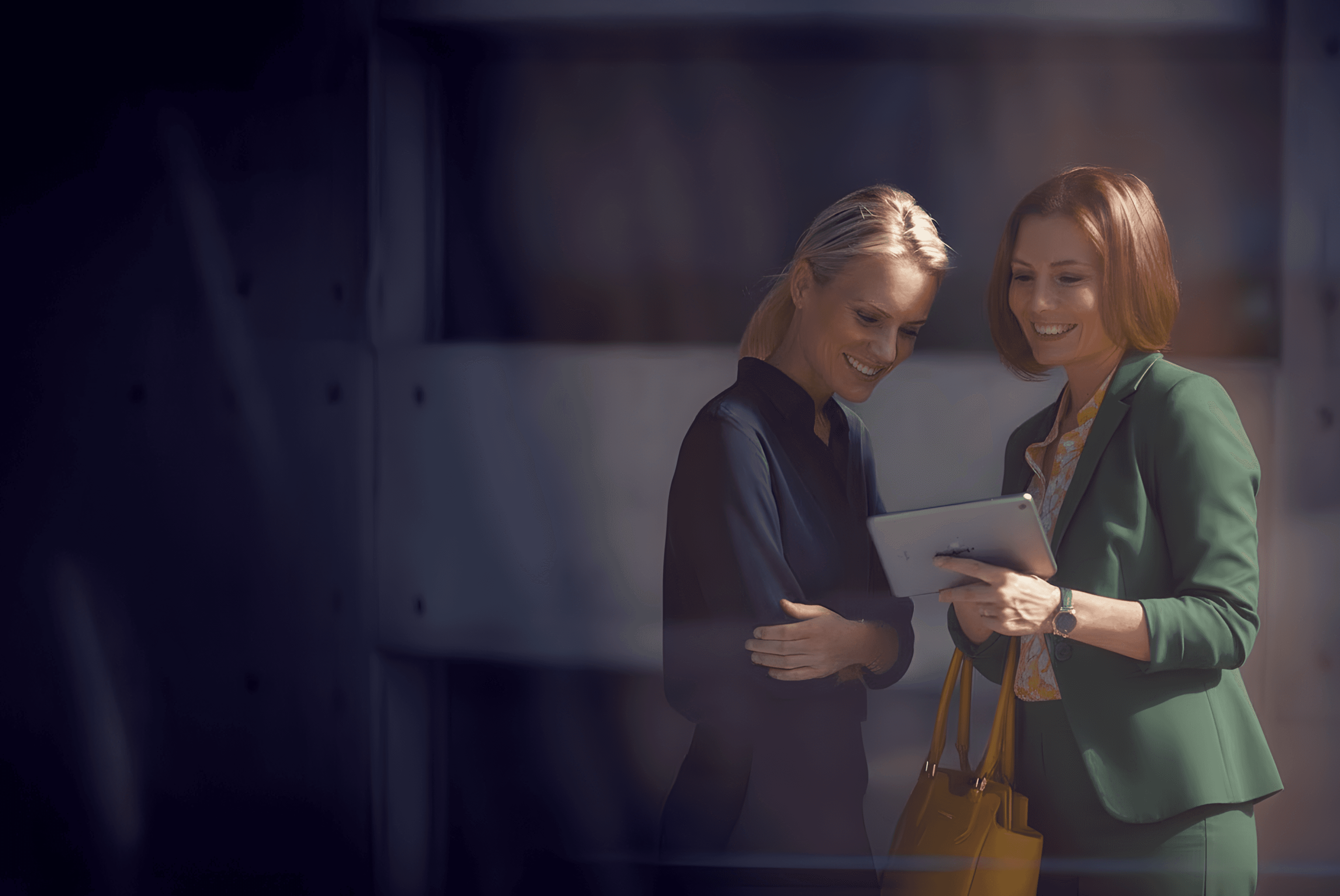 Channel partner - Two women looking at a tablet, smiling. One is wearing a green blazer and holding a yellow bag.