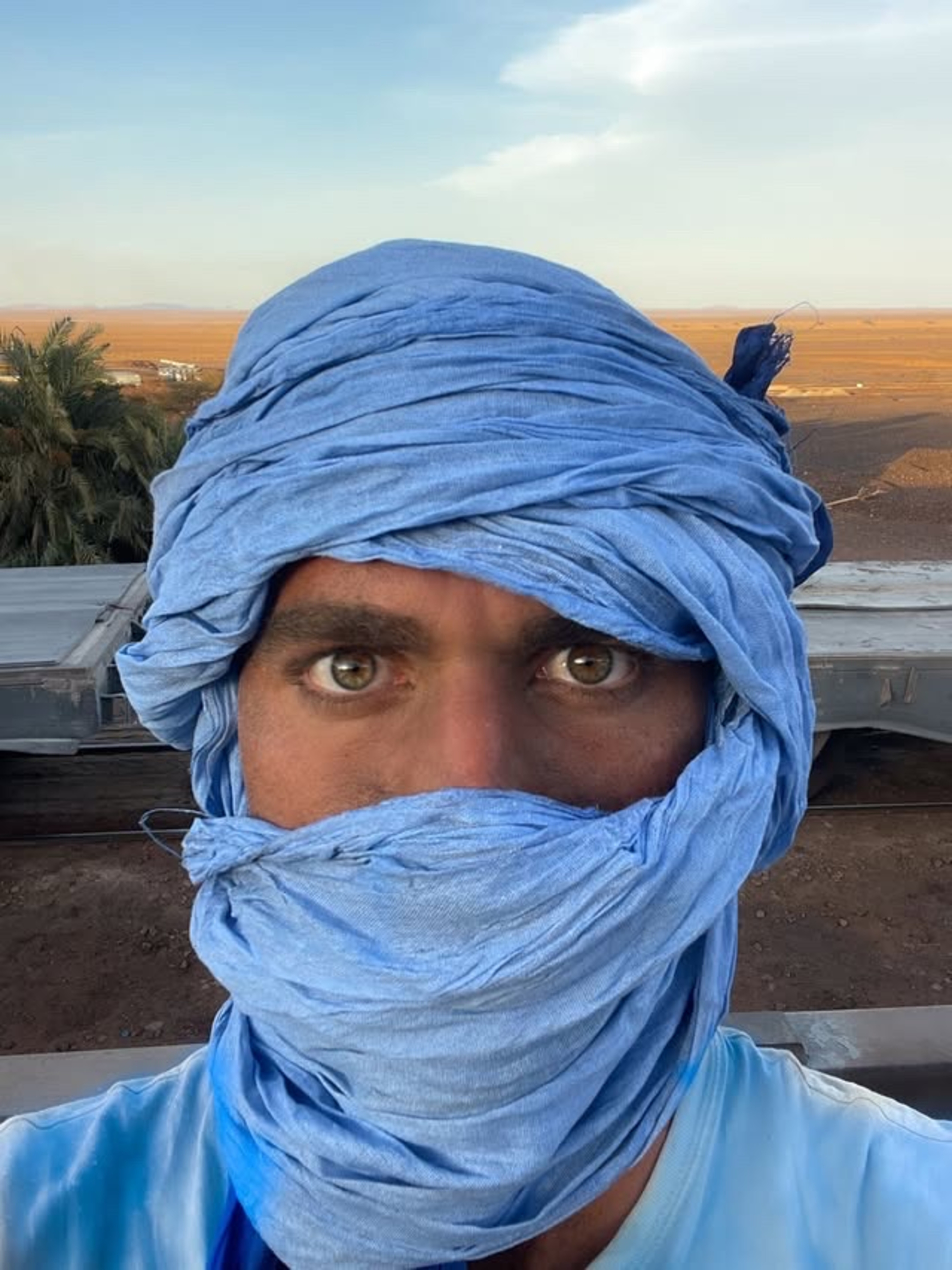 Close-up of a man's face in the desert, his face is wrapped in a headscarf so only his eyes are visible.