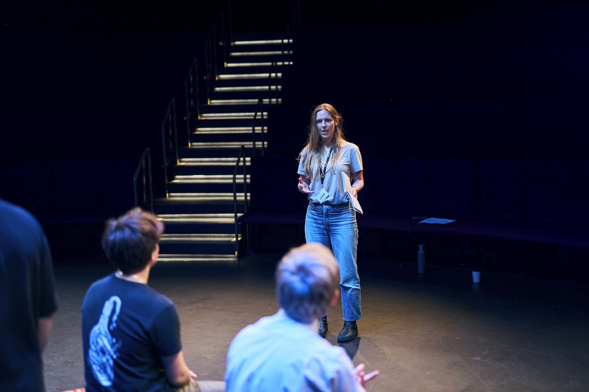 A facilitator or director stands in the centre of a theatre space with seating behind them, speaking to a small group of young people seated off-camera. They hold a script and gesture with their hands, appearing to lead a rehearsal or workshop.