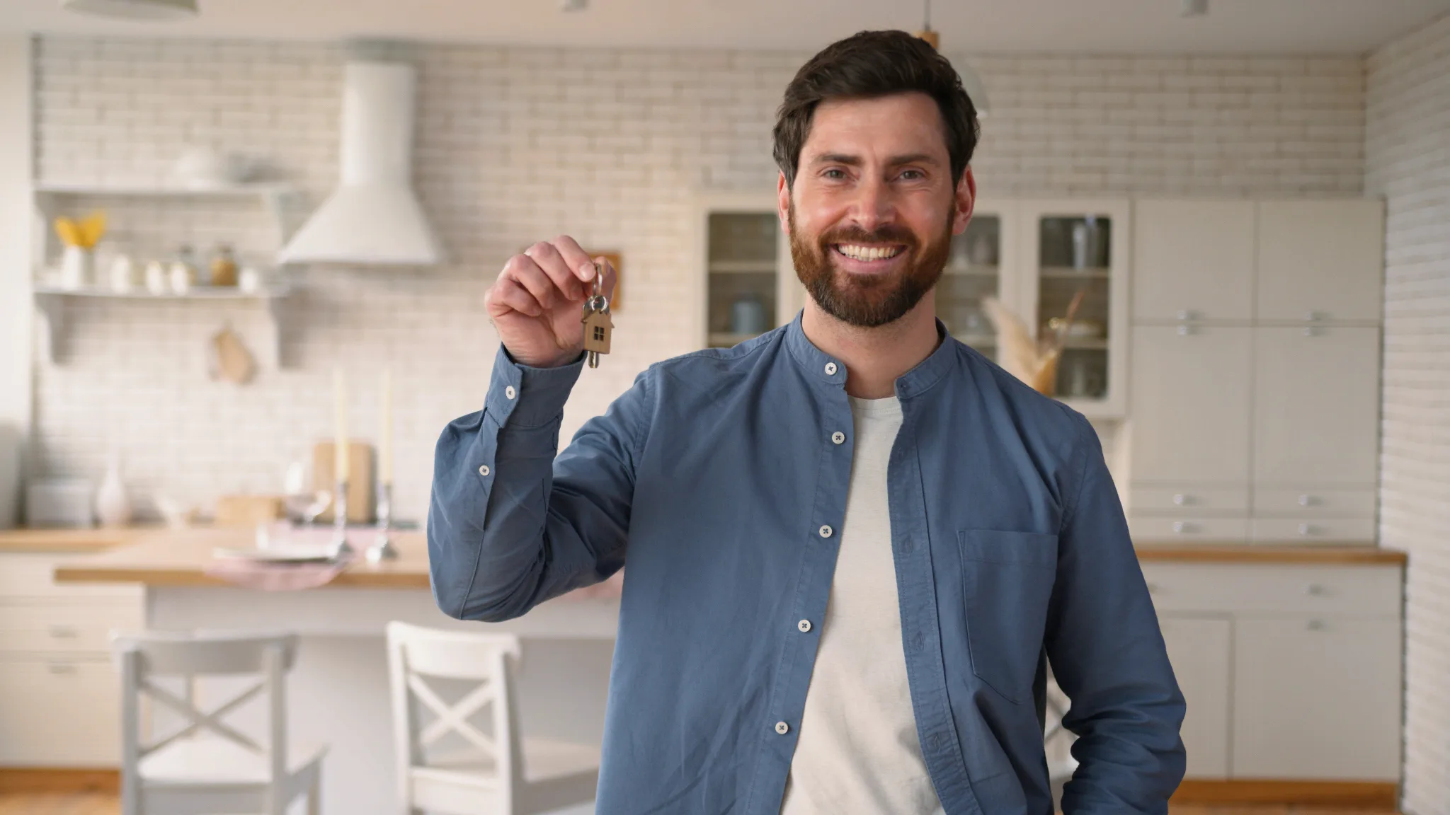 Bearded white man wearing a button down top, standing on the kitchen, holding house keys on one hand.
