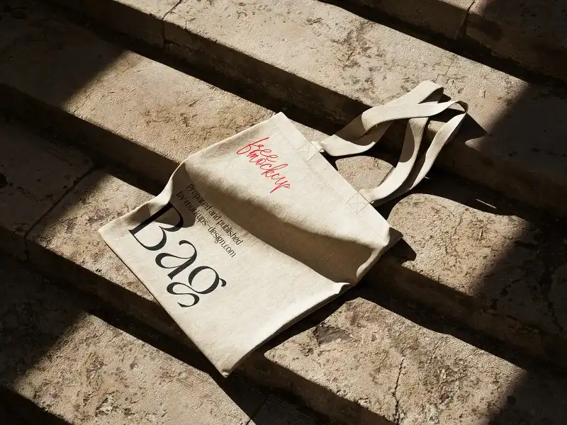 Natural fabric tote bag mockup placed on sunlit stone steps, with bold black and red text. The bag's texture and shadowing enhance its rustic, earthy appeal, perfect for eco-friendly branding presentations.
