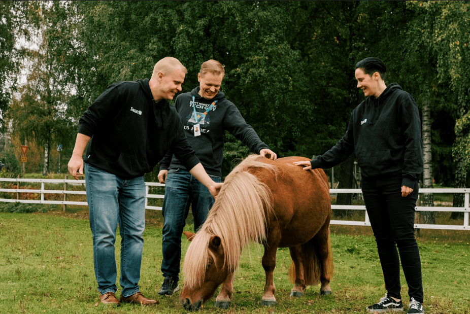 Three people smiling and petting a small brown pony in a grassy field