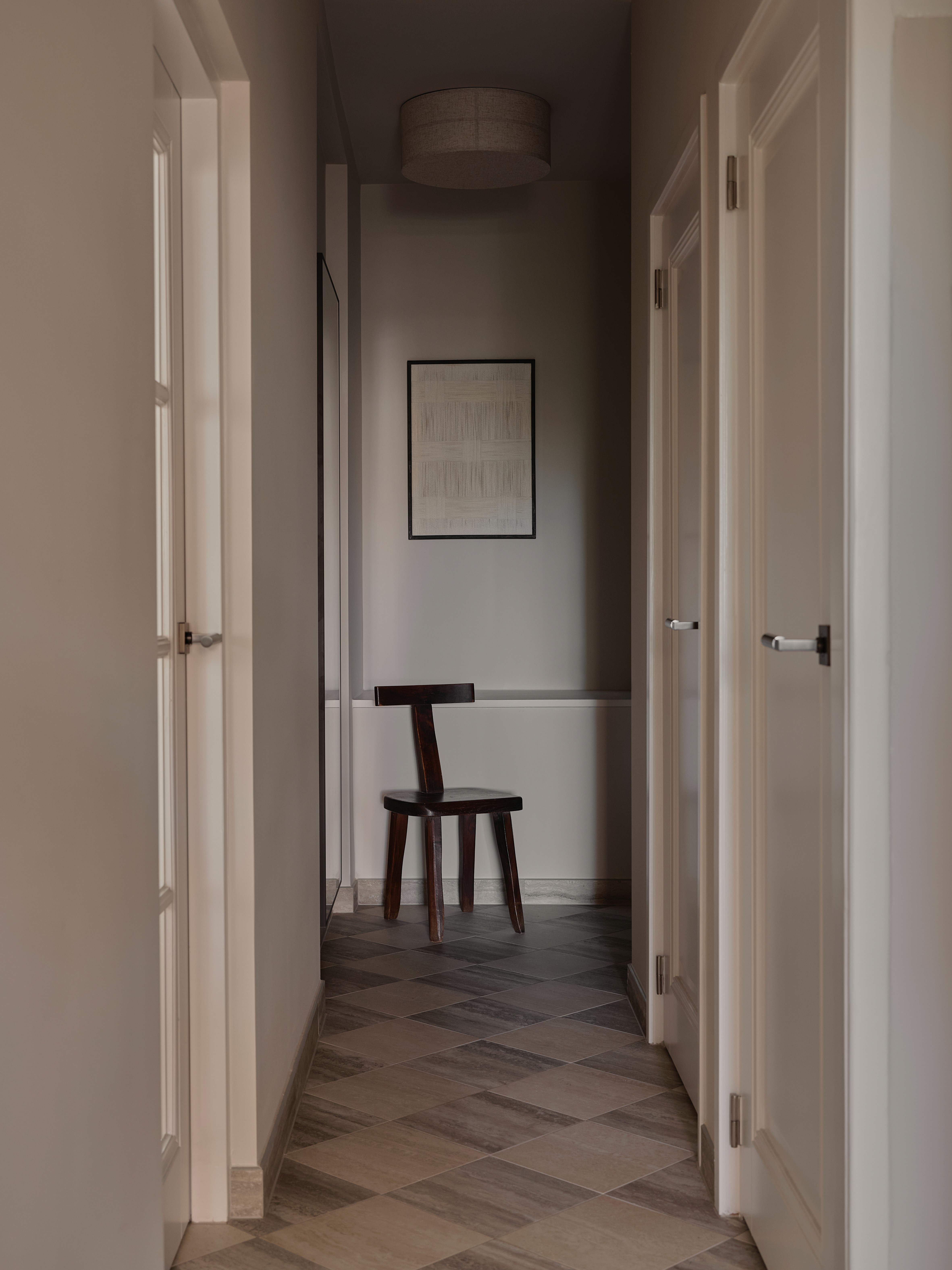 Hallway with white panelled doors, checkered stone tile floor, sculptural dark wood chair, abstract artwork, and linen drum ceiling light