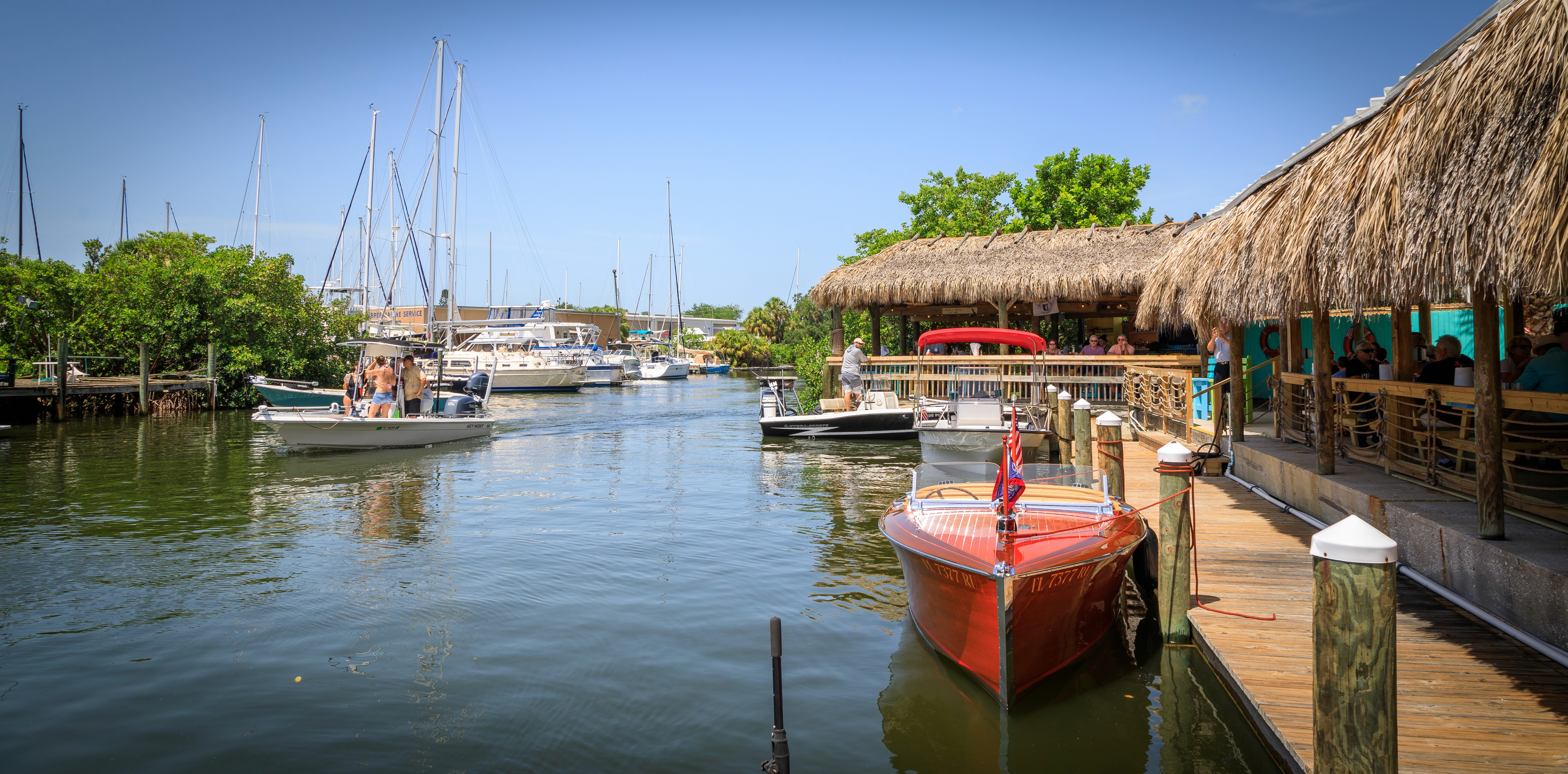 A serene marina scene featuring a rustic wooden dock with a thatched-roof restaurant on the right, several small boats moored and one red boat in the foreground, nestled under clear blue skies.