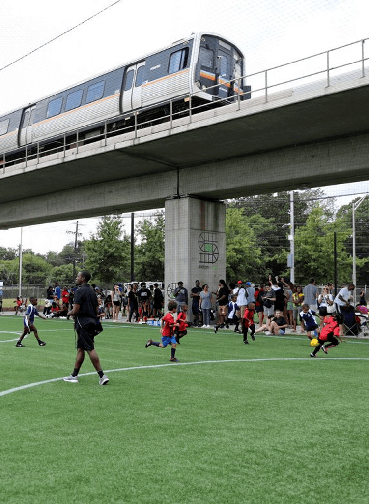 Children play soccer on a green field while a Marta train passes on an elevated track overhead. Spectators watch from the sidelines, and trees are visible in the background.