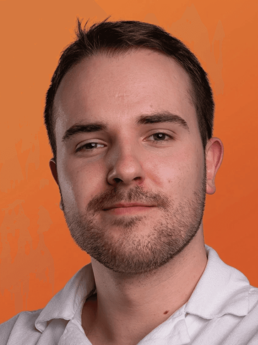 A professional, close-up studio portrait photograph of a young adult Caucasian man, approximately 30-35 years old, facing the camera. The man has short, dark brown hair, a neatly trimmed full beard, and a moustache. He is looking directly into the lens with a neutral, slightly friendly expression and hazel-brown eyes. He is wearing a simple white cotton polo shirt with a slightly textured fabric and a visible collar. The composition is a head and shoulders shot, centered within the frame. Notable facial features include a slight, healing mark on his forehead and visible skin texture with some redness and a mole on his lower neck. The background is a seamless, vibrant, solid orange color, evenly and brightly lit, providing a sharp contrast. The lighting is soft and flattering studio light, illuminating his face without harsh shadows. The image is a standard professional profile or headshot.