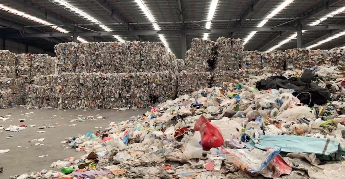 Baled and loose waste piled inside a large sorting facility, with plastic bags and packaging across the floor