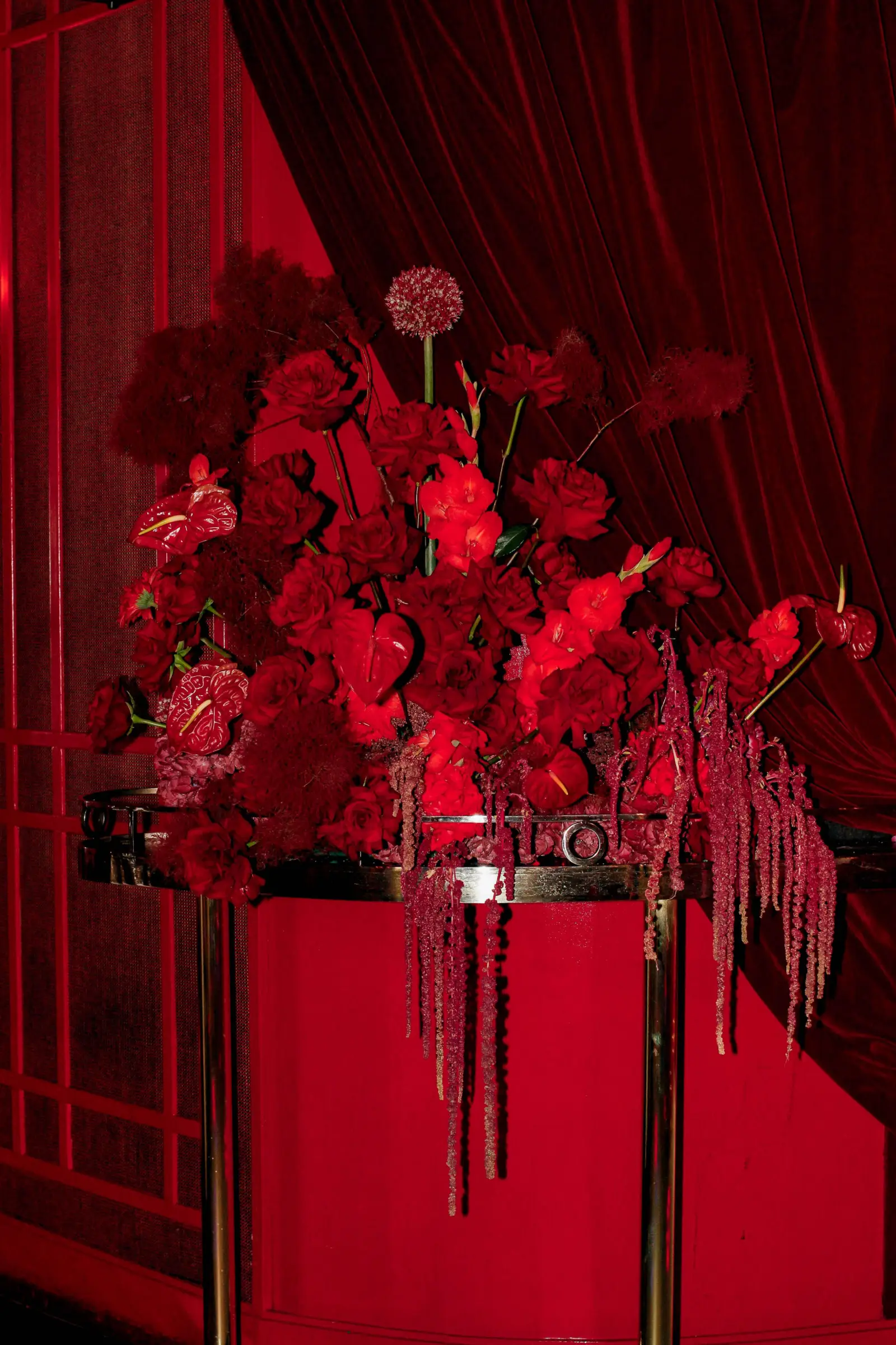Cascada floral de rosas y amaranto rojo sobre mesa dorada frente a cortinas de terciopelo carmesí.