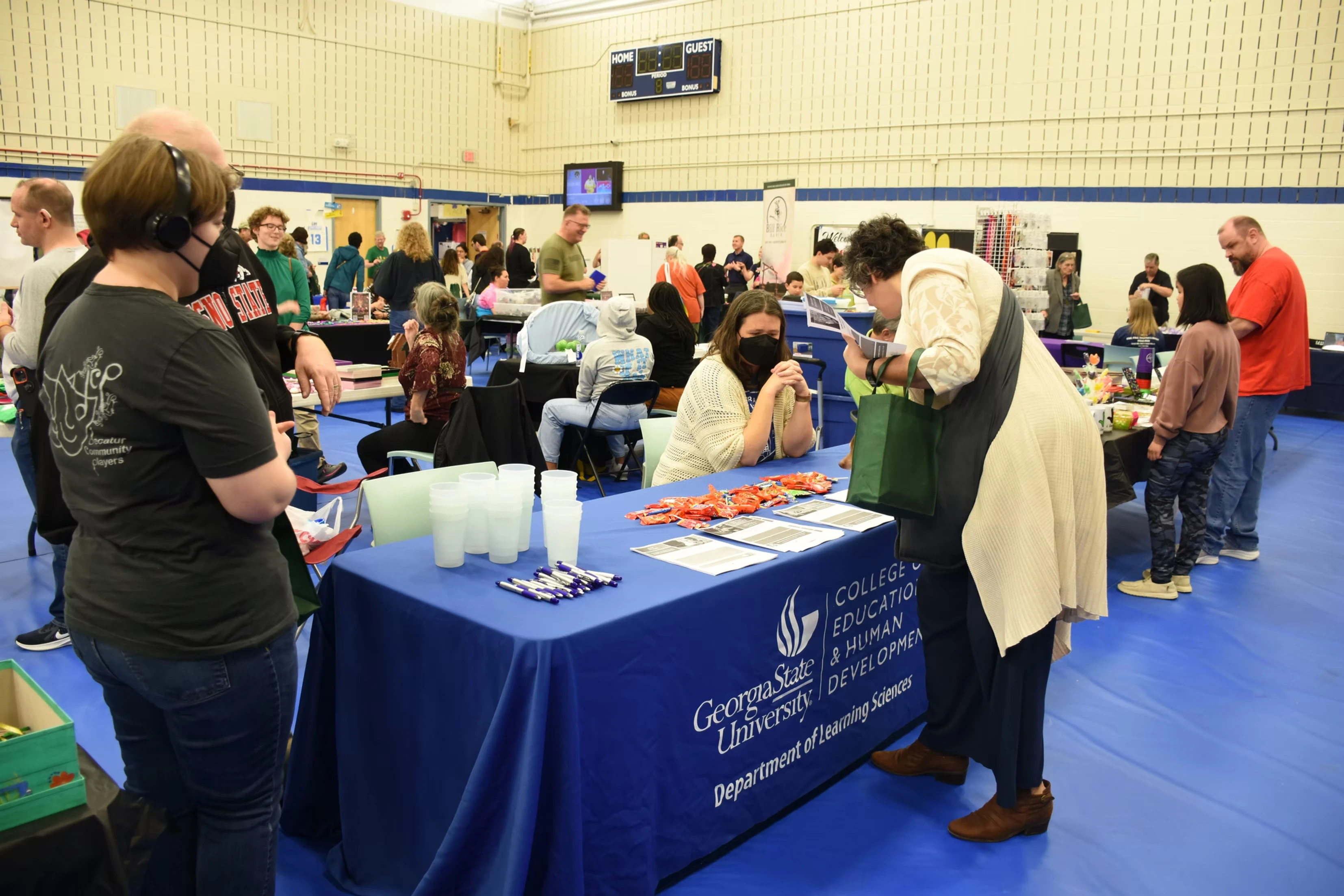 A college fair in a gymnasium. People browse tables with information, some wearing masks. A table displays the George Mason University logo.
