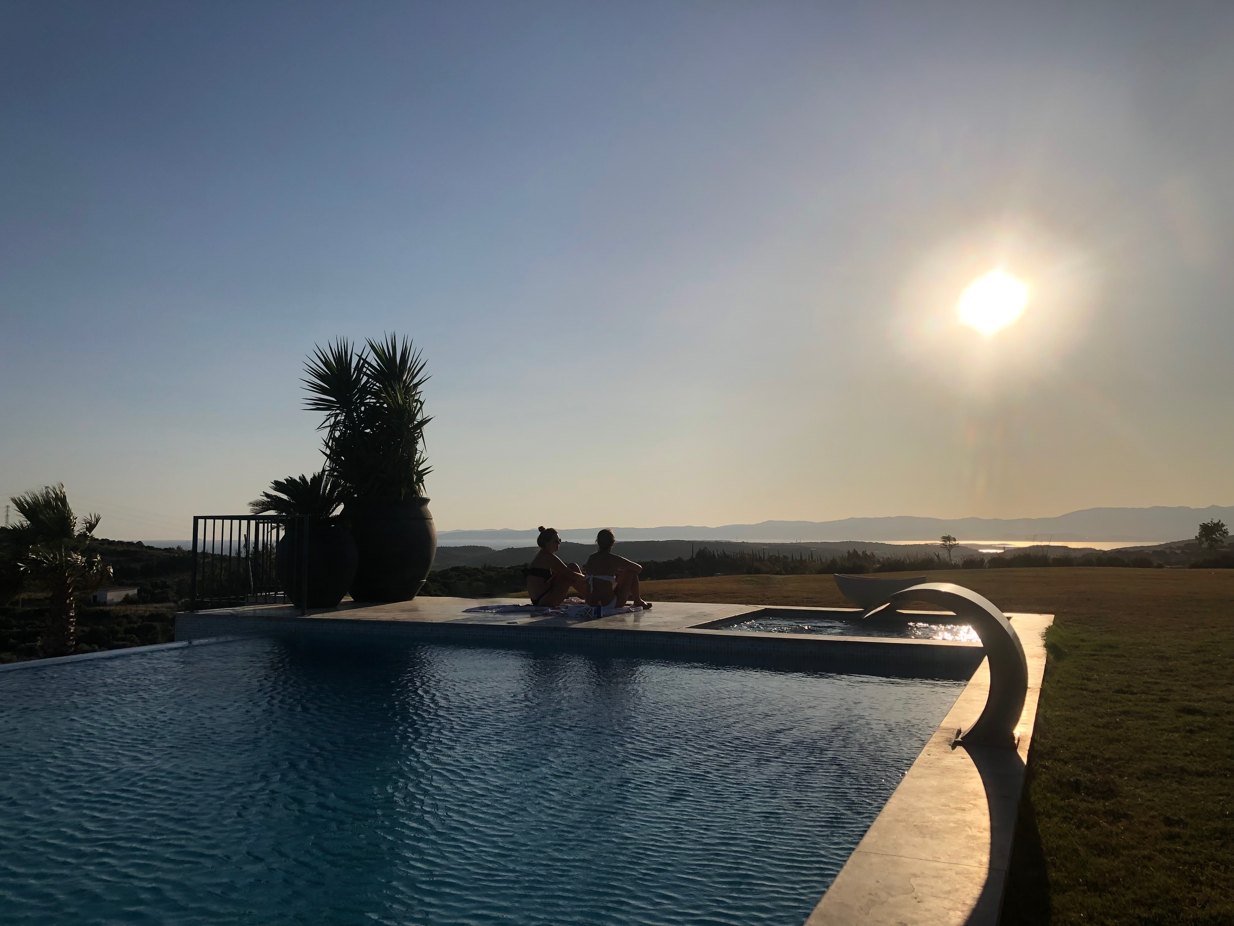 Two women sitting cross-legged in silhouette on the pool terrace at Villa Laluli, watching the sunset over the Aegean landscape in Alaçatı, Turkey.