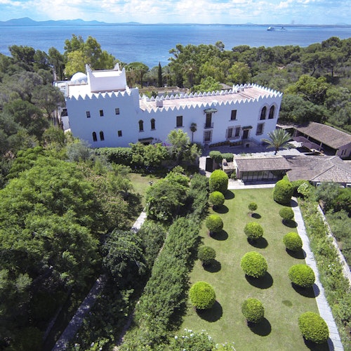 Aerial view of a white castle-like building with a manicured garden, surrounded by lush greenery, near the sea.