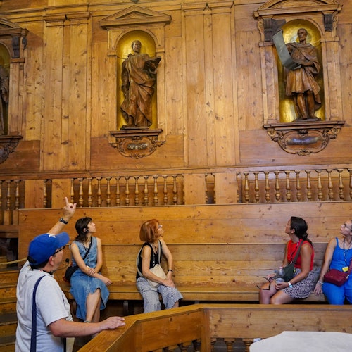 ארבעה אנשים יושבים על ספסלים מעץ בחדר עם קירות מעץ ופסלים דתיים. המדריך מצביע לעבר הפסלים.