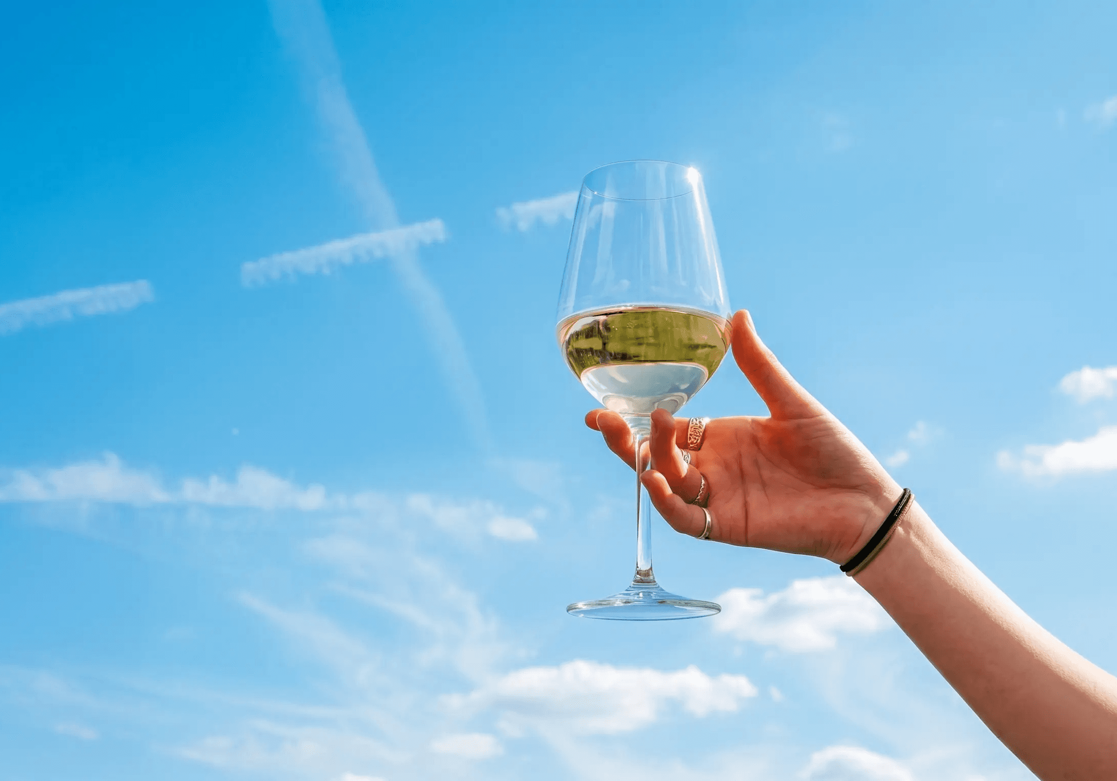 Main tenant un verre de vin blanc devant un ciel bleu.