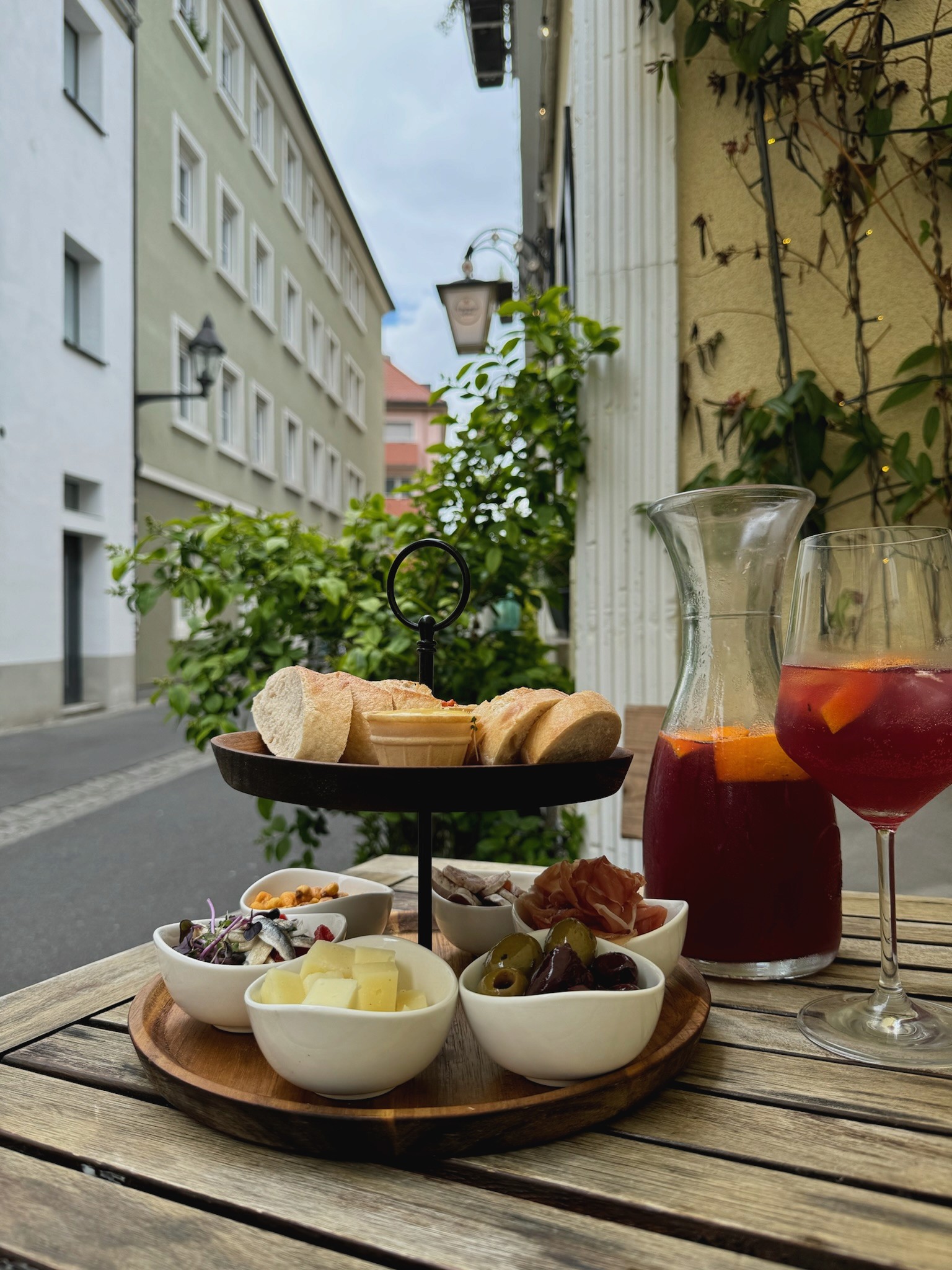 Holztablett mit verschiedenen Antipasti, Brot und einer Karaffe Sangria auf einem Holztisch im Freien, im Hintergrund eine Straße und Gebäude.