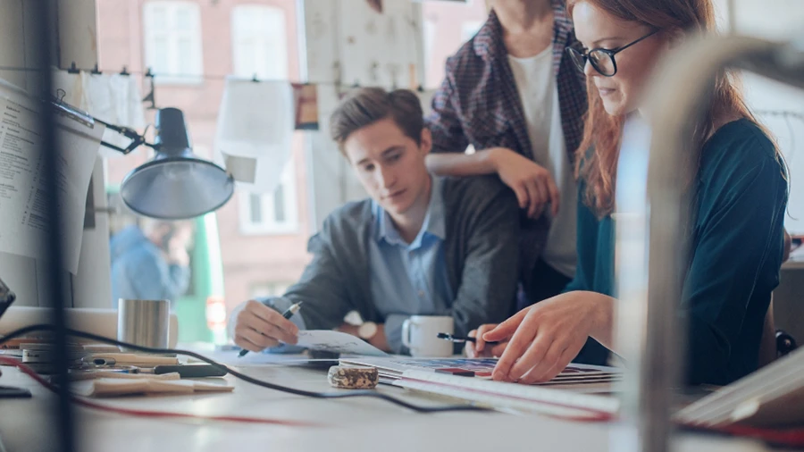 Group of people collaborating at a desk, reviewing documents and designs in a bright office.