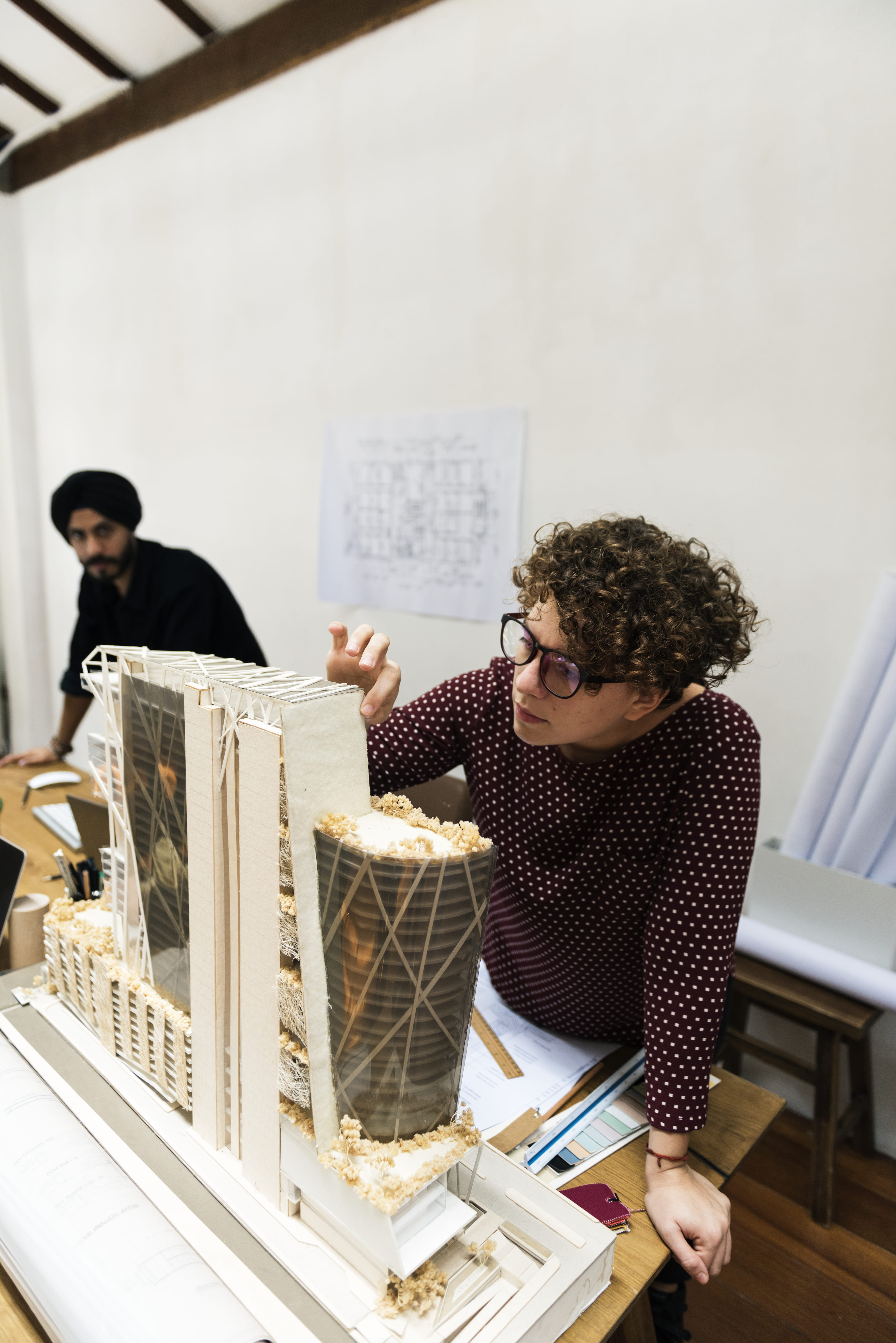 A woman examines a detailed architectural model of modern skyscrapers on a wooden table, with blueprints and laptops nearby in a well-lit room.