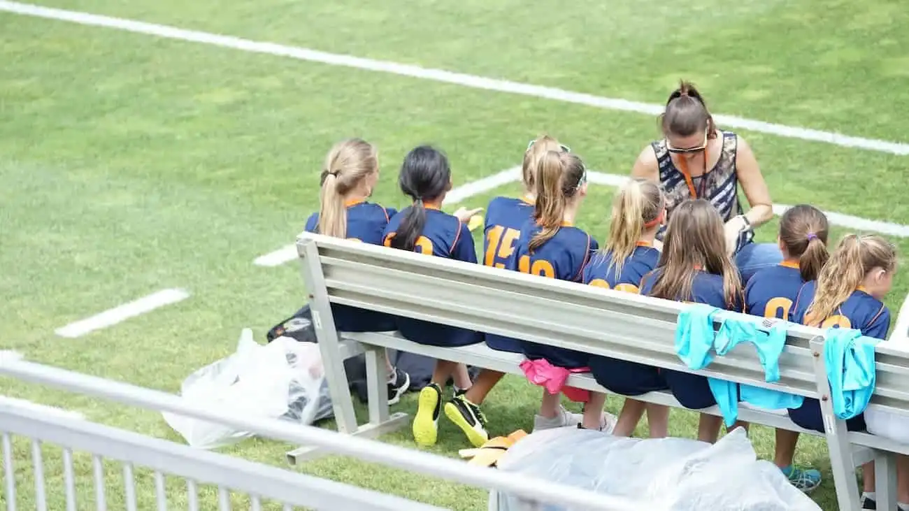 In this image, a coach is seen giving tactical instructions in the coaching zone, surrounded by young football players sitting on a bench. This represents the important role of the coaching zone, where communication between coach and players takes place, and emphasises the importance of the technical zone in modern football.