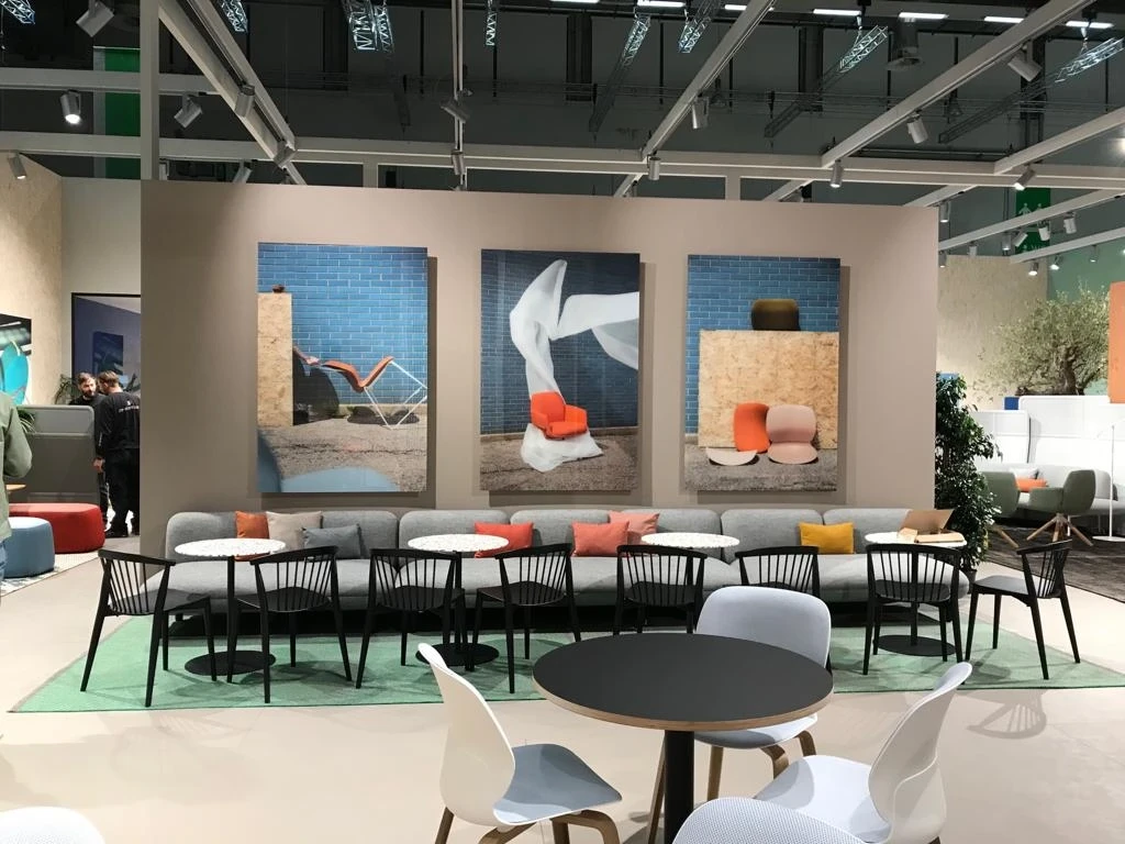 Lounge at the HAWORTH ORGATEC stand with a long grey sofa, round bistro tables, black wooden chairs and three large furniture campaign prints on the wall.