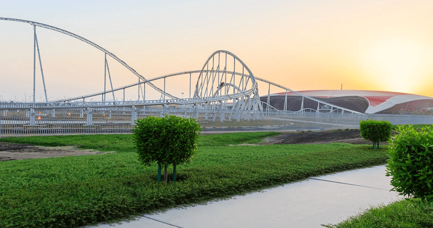 A roller coaster silhouetted against a vibrant sunset, with a lush green field in the foreground.