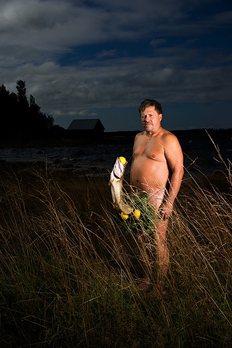 A man stands shirtless in tall grass at night, holding a fish with yellow fins and tail in his left hand. He looks directly at the camera with a neutral expression. Behind him, a body of water and a small building are visible under a dark sky with scattered clouds.