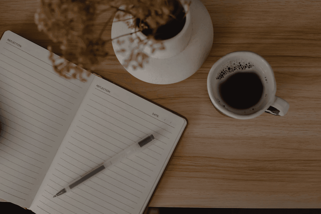 A cup of coffee next to a notebook and a pen on a wooden table.