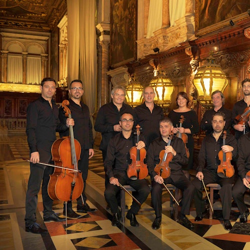 A group of musicians dressed in black, holding string instruments, posed in an ornate hall with chandeliers and paintings.