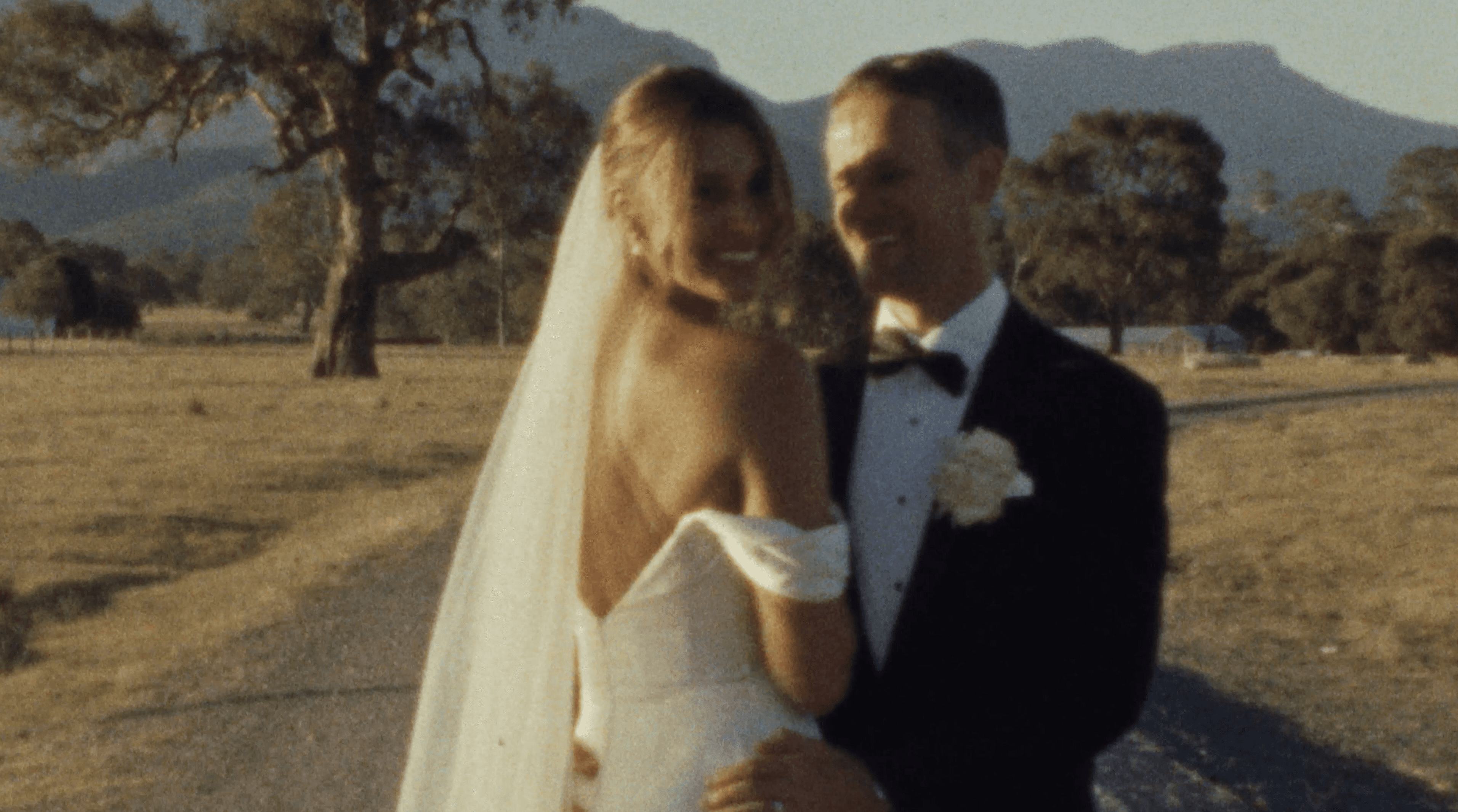 A joyful bride and groom, dressed in an elegant white gown and classic black tuxedo, celebrate their wedding outdoors, surrounded by falling white rose petals and lush greenery.