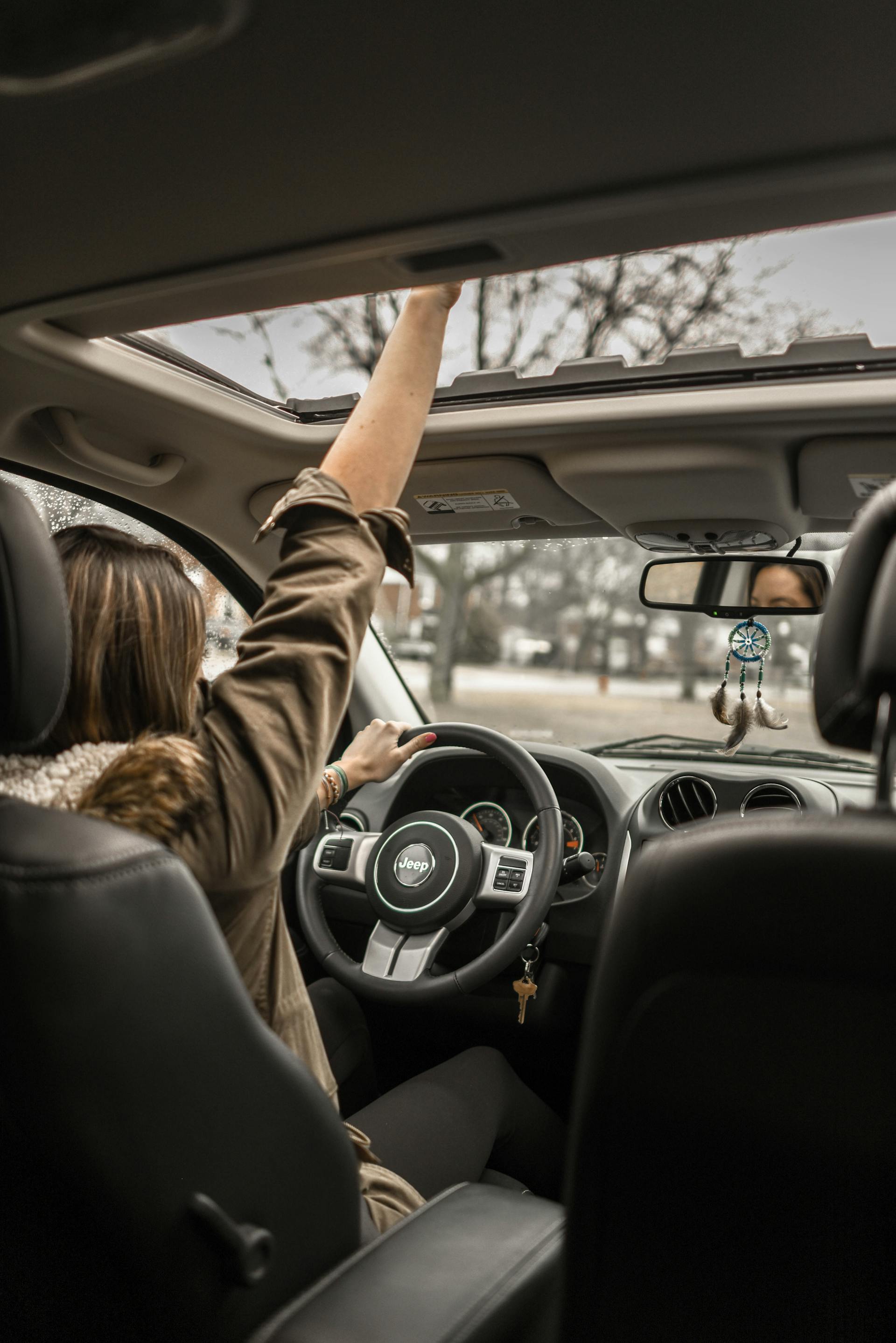 A vertical shot from the back seat of a car showing a woman driving. She is wearing a brown jacket, and her right hand is raised toward the open sunroof. The steering wheel has a Jeep logo. The road and trees are visible through the windshield.