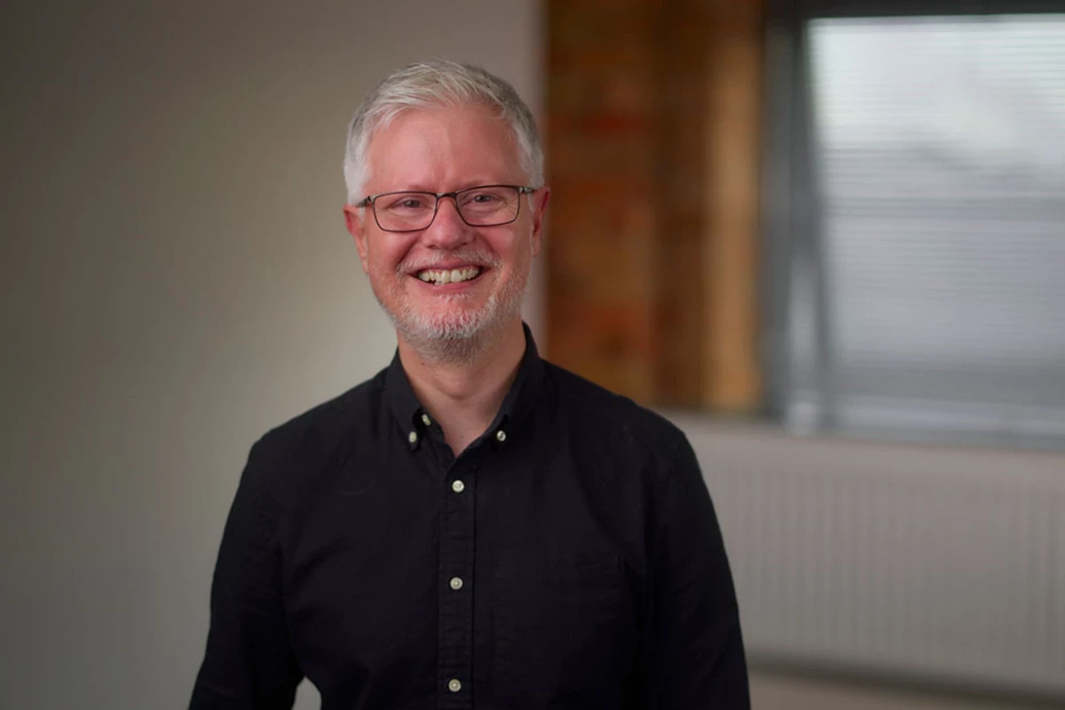 A man with grey hair and glasses wearing a black shirt smiles at the camera, a white wall with a window and an orange curtain is in the background