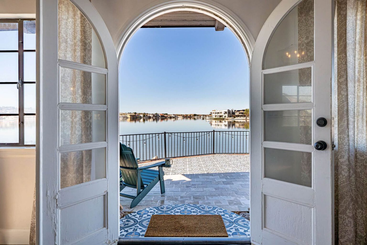 Arched doorway opening to a backyard patio overlooking a lake, with a teal-blue chair and Spanish-style tile flooring.