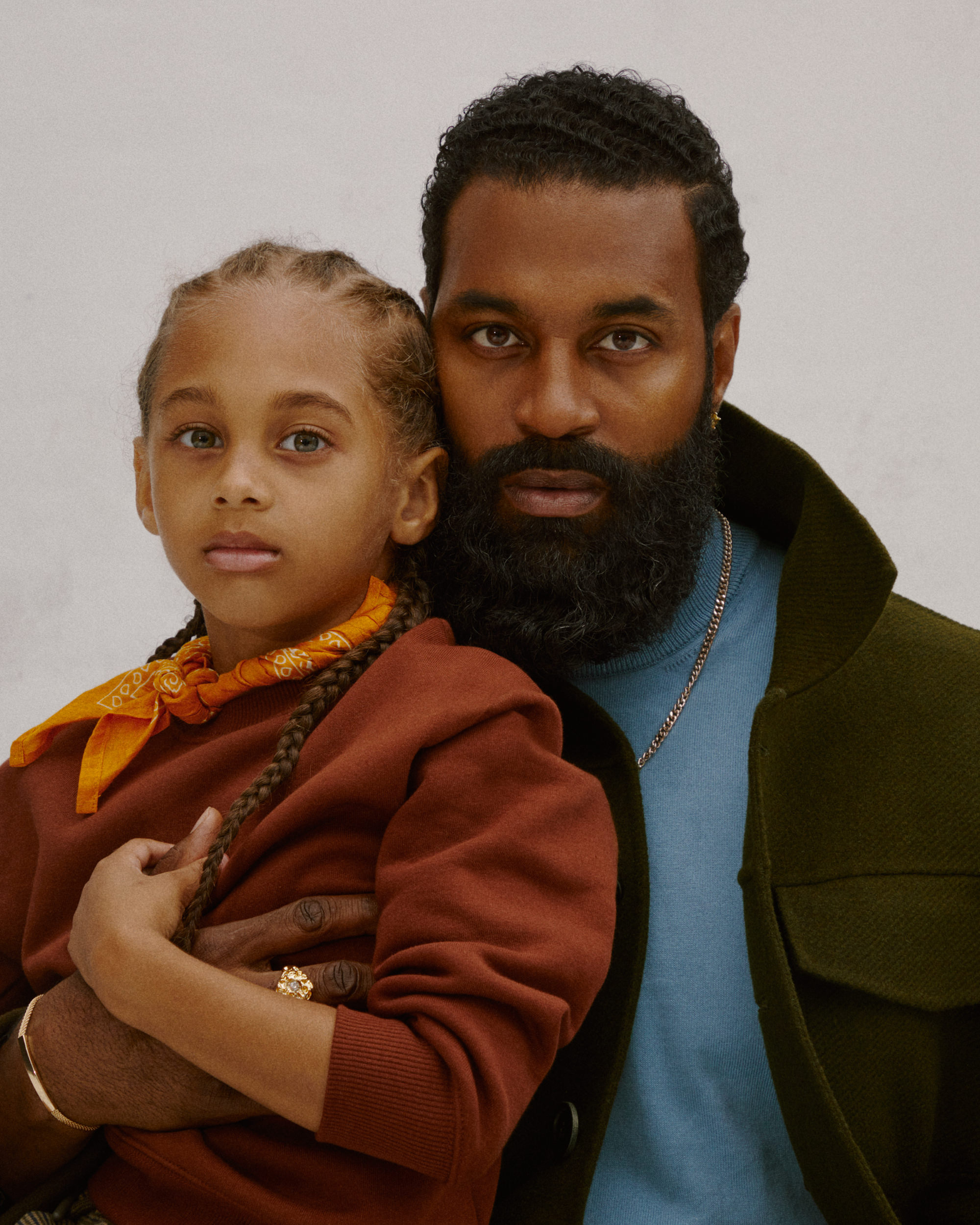 A father holds his young son close, both dressed in warm tones, his head resting near his chest.