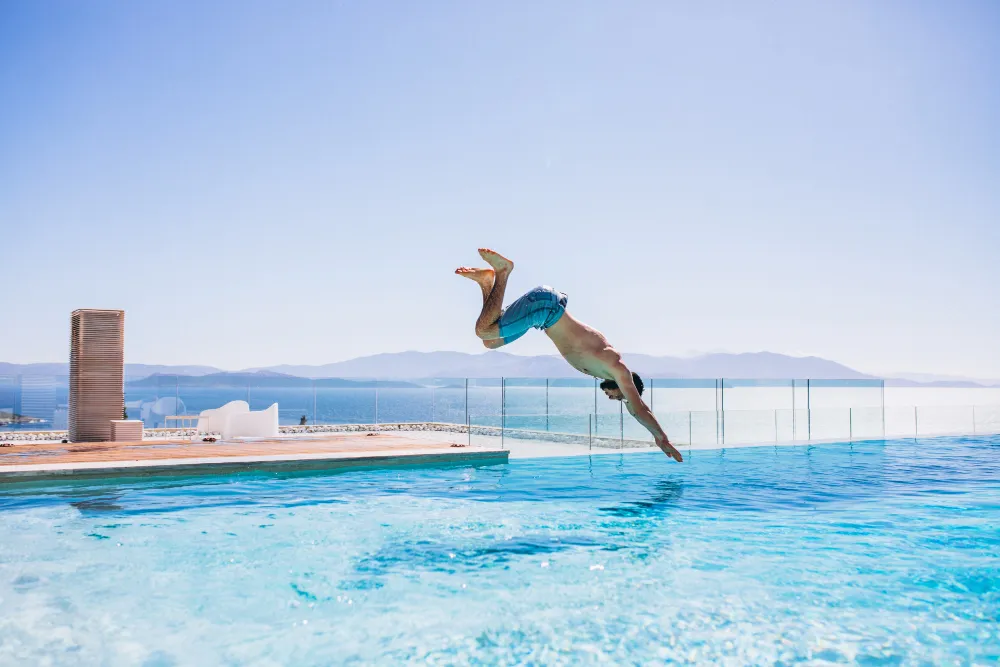 Man diving into a bright blue outdoor pool with mountains and the ocean visible in the background.