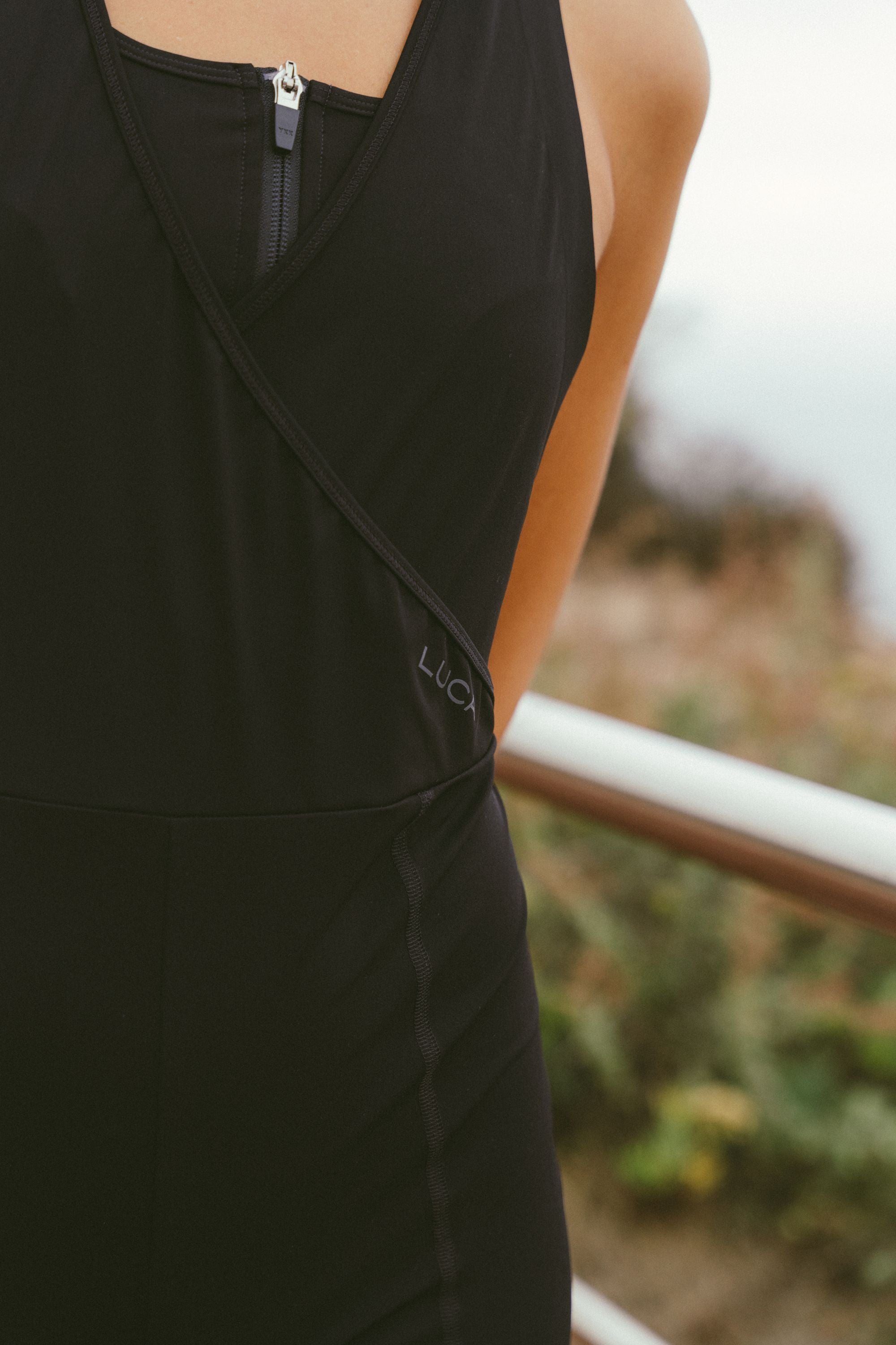 Extreme-close-up-shot-of-the-torso- area-of-a-black-athletic-skinsuit- trisuit-The-garment-features-a-black-on-black-embroidered-logo- reading-"LUCA"-near-the-waist-hip- below-a-cross-over-neckline-with-a- visible-silver-zipper-The-background-is-blurred-outdoors-with-foliage-and-a-railing