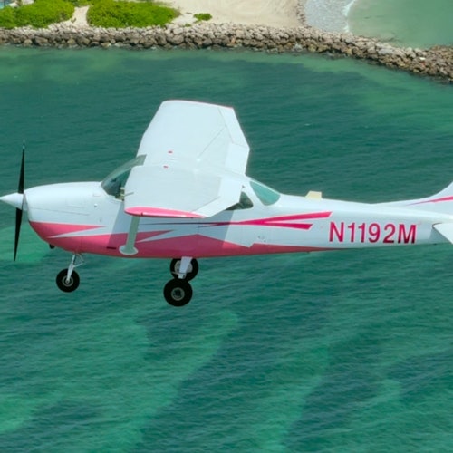 一架小型粉白相间的飞机在清澈碧绿的海水上空飞行，靠近岩石岸边。