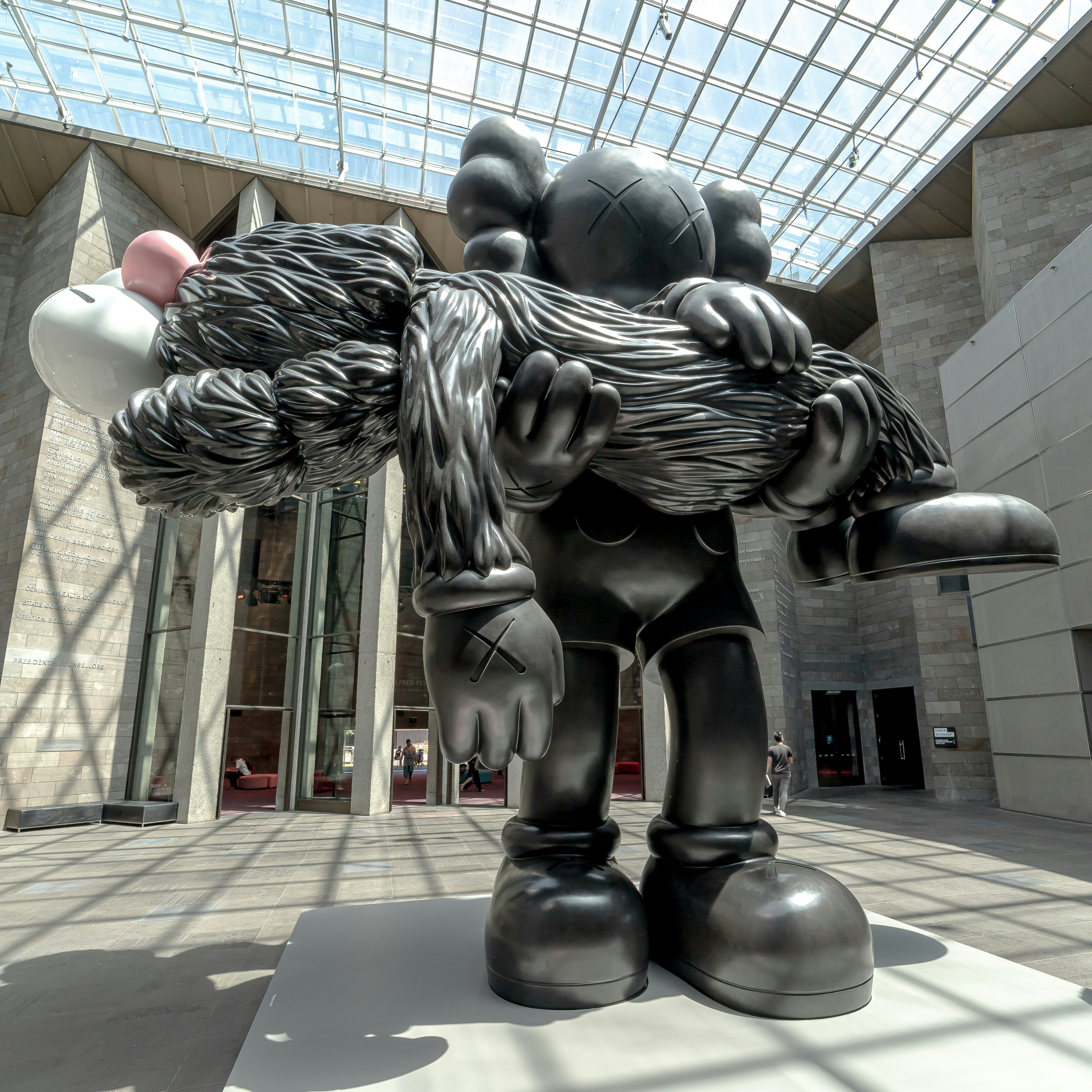 A large-scale black sculpture by KAWS titled 'GONE,' featuring the iconic Companion character carrying a limp, furry BFF figure. The piece is exhibited in a bright, modern museum atrium with a glass ceiling, illustrating the intersection of street art iconography and contemporary fine art.