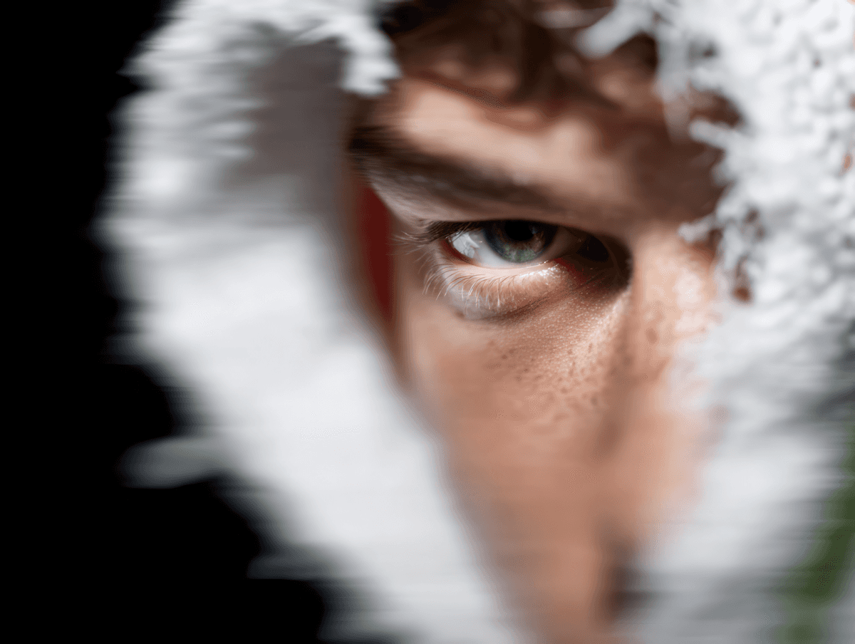 Close-up of a green-eyed person framed by a white textured material, with dramatic lighting emphasizing their eye and skin texture.