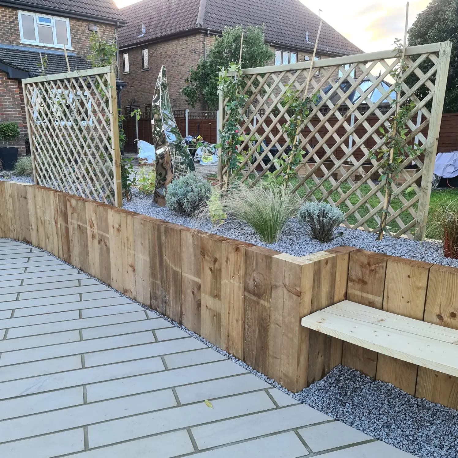 A wooden garden structure features lattice panels, planters, and a bench, set in a landscaped area.