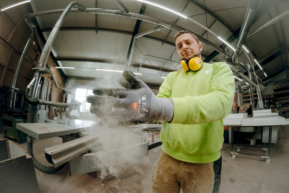 Ein Mann in einer Werkstatt lässt Staub aus seinen Händen fallen. Er trägt Handschuhe, Ohrenschützer und einen grünen Pullover.