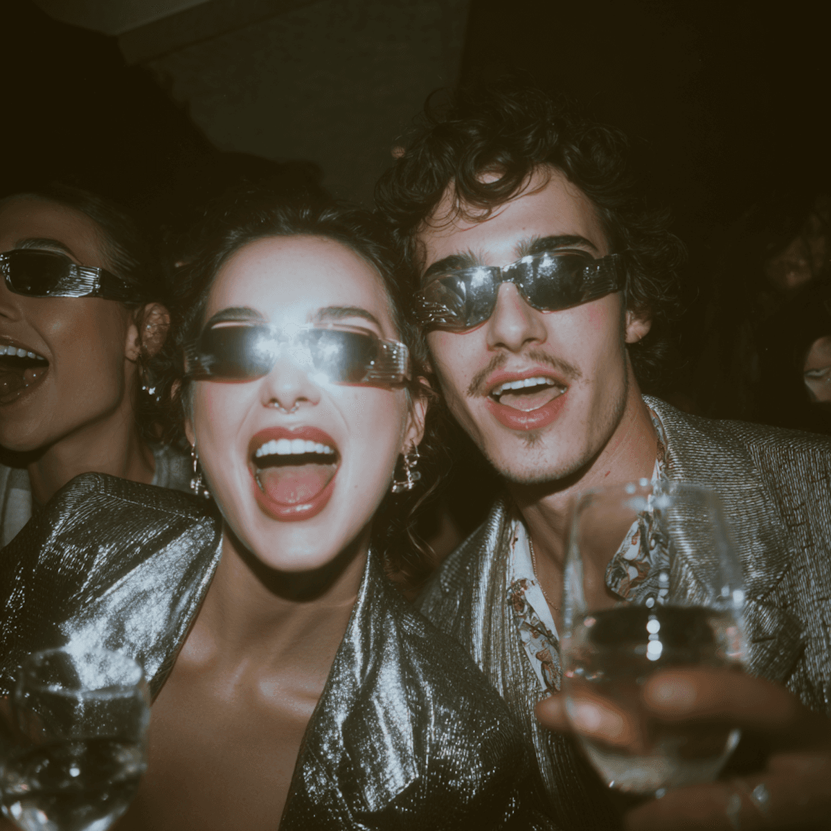 Man and woman wearing sunglasses and laughing while holding a glass of wine at a reception.