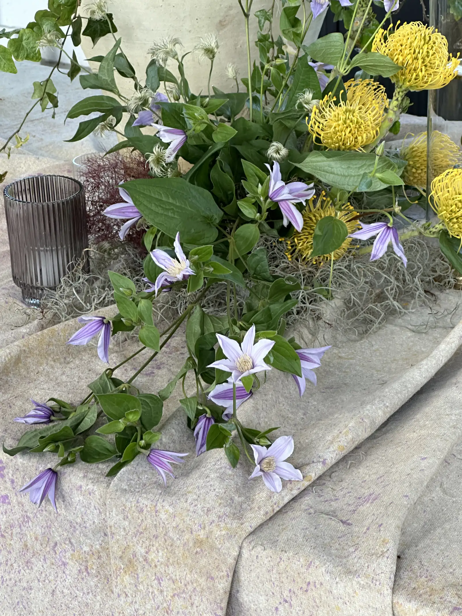 Arreglo floral con clematis lilas y proteas amarillas sobre un mantel texturizado, acompañado de follaje verde y un vaso de vidrio estriado con vela.