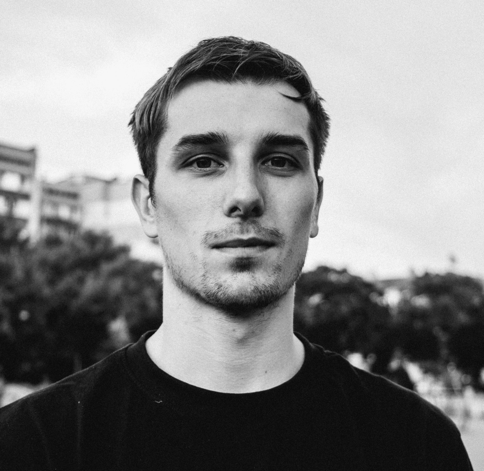Black-and-white portrait of a young man with short hair and light facial stubble, looking directly at the camera with a neutral expression, outdoors with blurred buildings and trees in the background.