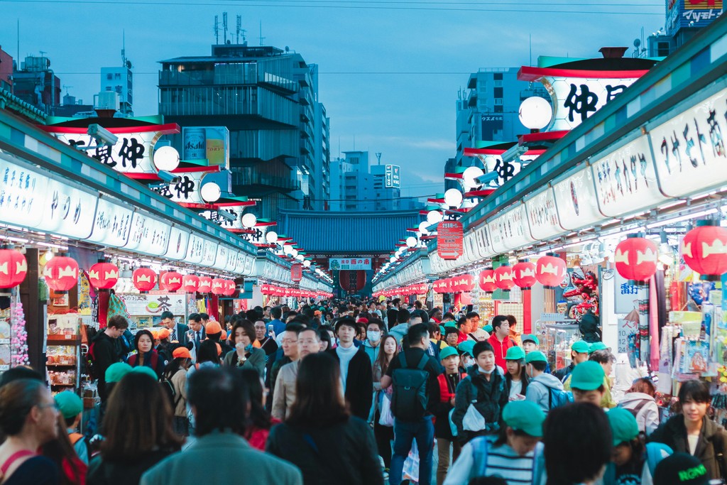 A bustling outdoor market in Tokyo, Japan, lined with brightly lit stalls displaying Japanese signs and lanterns, filled with locals and tourists exploring the vibrant array of goods and street food.