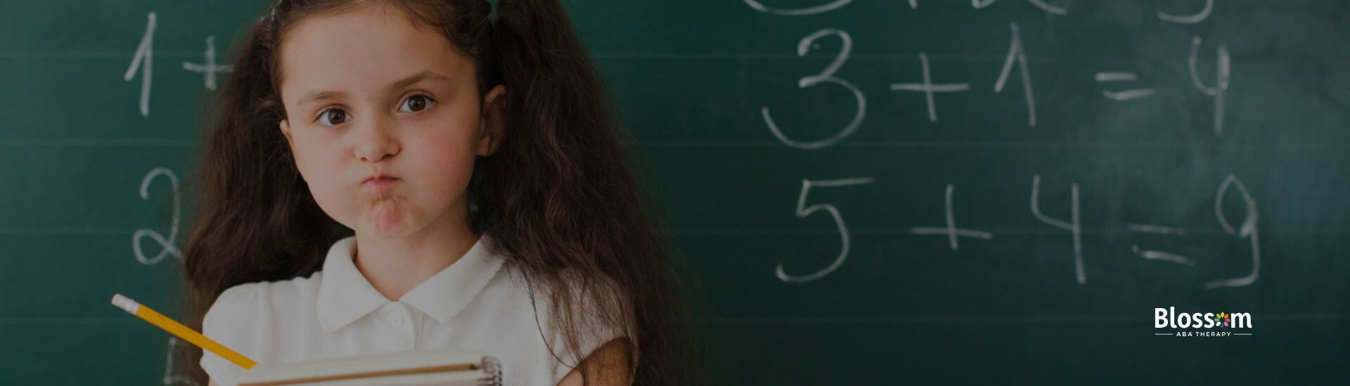 Young girl with autism looking frustrated while holding a notebook in front of a math chalkboard.