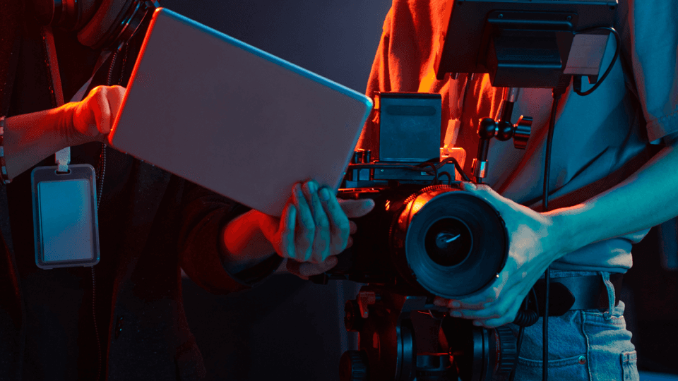 A young person wearing a tan hoodie sits at a desktop computer, focused on the screen while pointing toward something displayed. An older adult wearing glasses and a dark jacket leans in beside them, looking at the monitor and appearing to provide guidance. The setting appears to be a modern classroom or workspace, with another person working at a computer in the background.