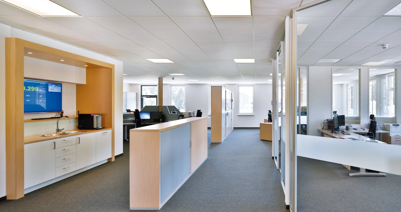 Modernes Büro mit hellen Arbeitsplätzen, Glaswänden, Küchenzeile und einem Bildschirm mit Zahlenanzeige.
