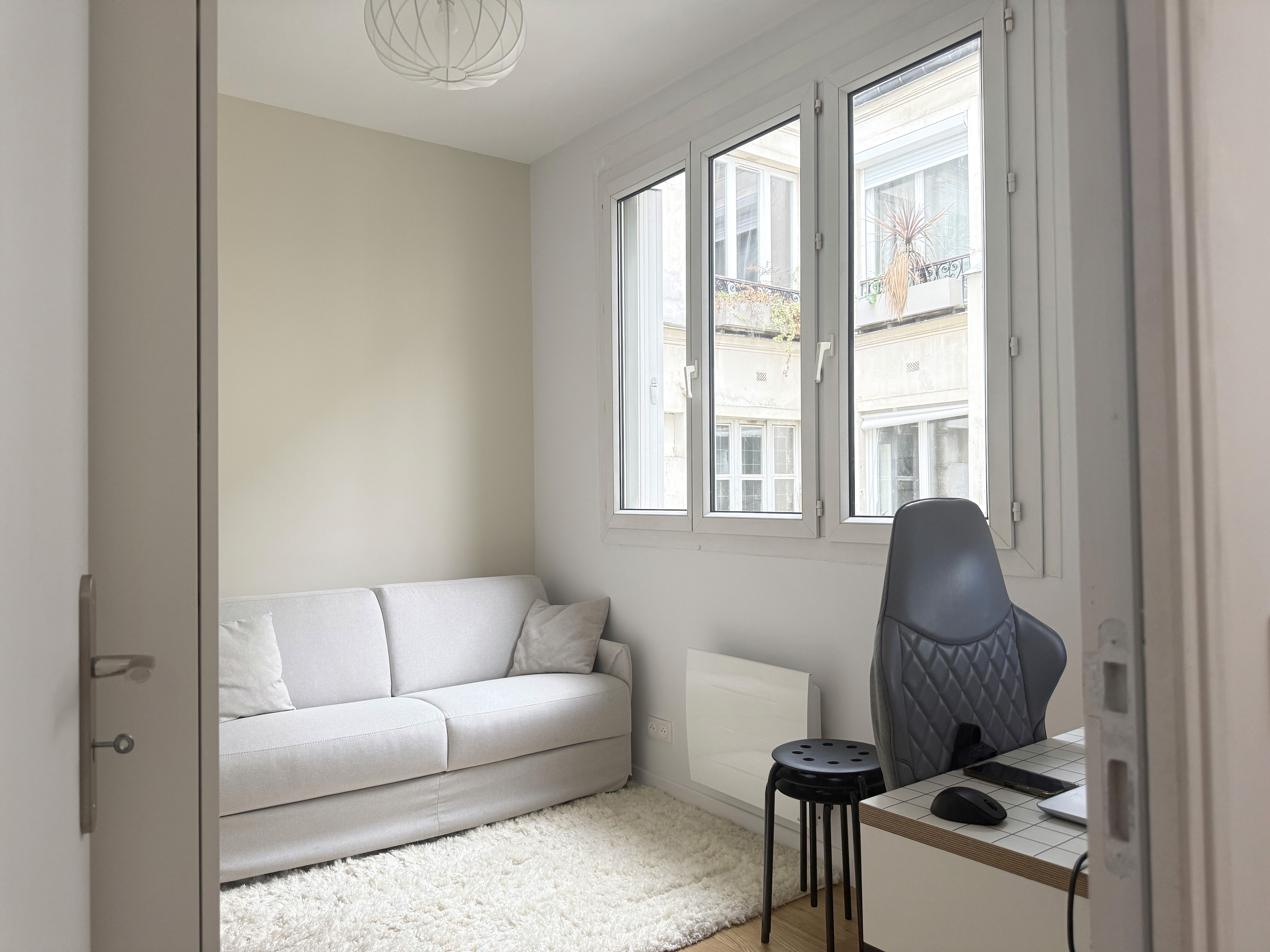 Deuxième chambre aménagée avec un canapé gris clair sous de grandes fenêtres, créant un espace de détente lumineux.