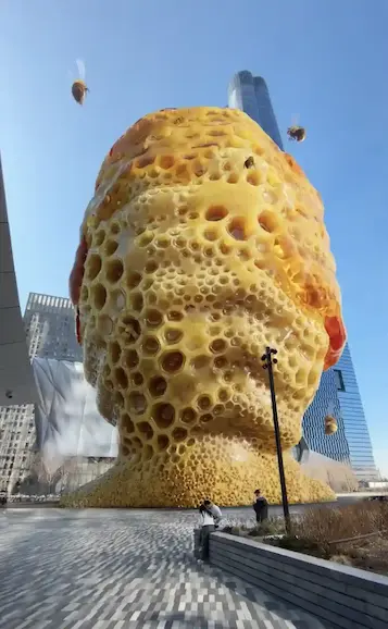 A massive golden human head sculpture made of honeycomb cells, with bees flying, set in a modern city plaza.