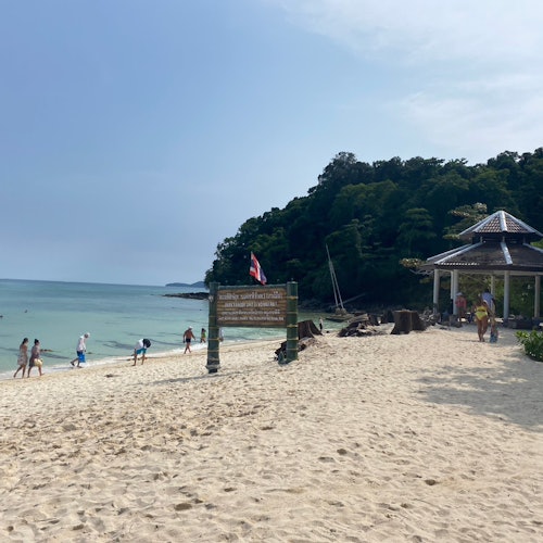 A beach scene with people walking on the sand, a wooden sign, a pavilion, and a forested hill in the background.