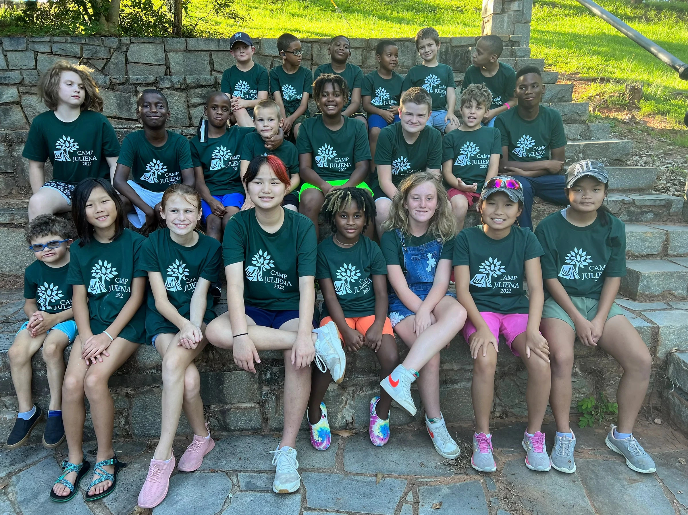 Group photo of campers in green Camp Juliena t-shirts sitting on stone steps outdoors.